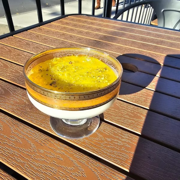 A golden-rimmed glass bowl of yellow Afghan dessert with nuts sits on a wooden outdoor table.